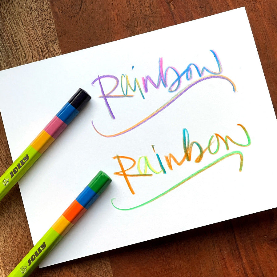 Jolly Rainbow pencils set on a white note card with the words "Rainbow" written twice showing the rainbow swirl writing and a drawn line below set on a wooden table