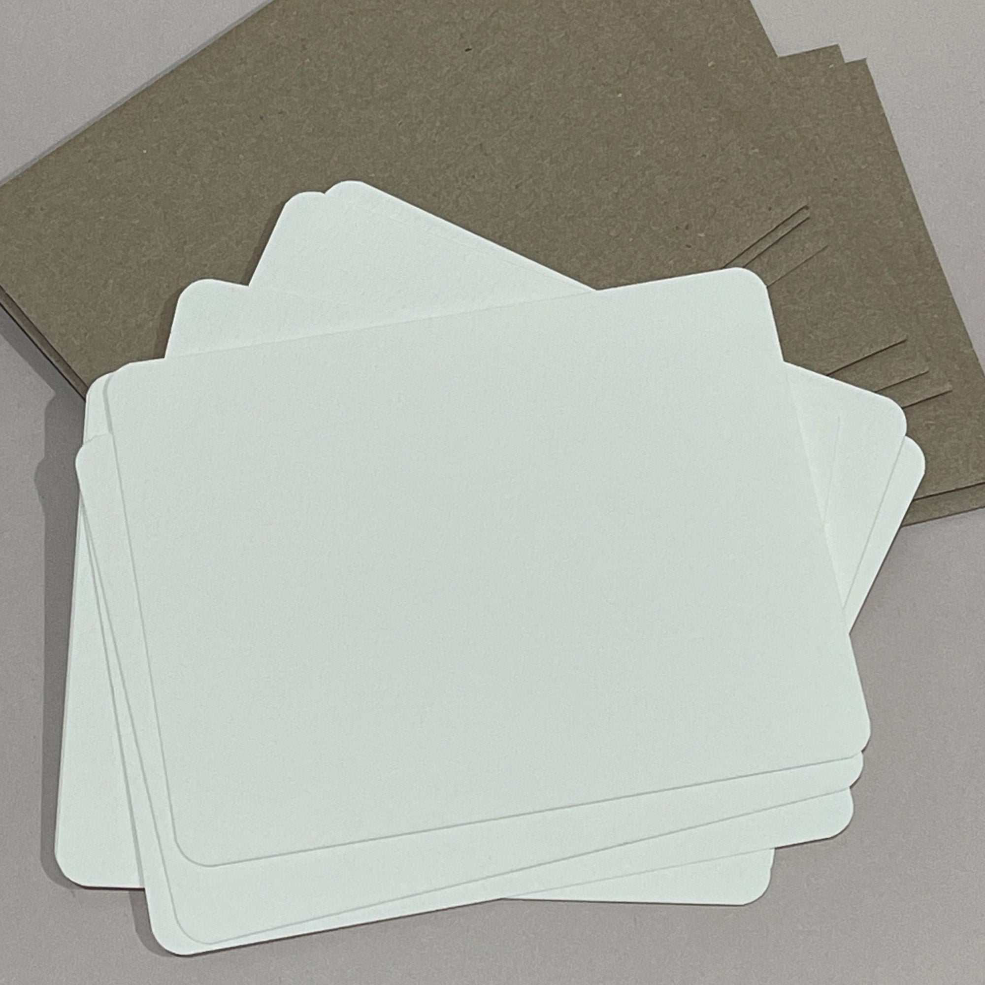 A stack of blank watercolor paper flat cards with rounded corners on top of a stack of kraft paper A2 envelopes
