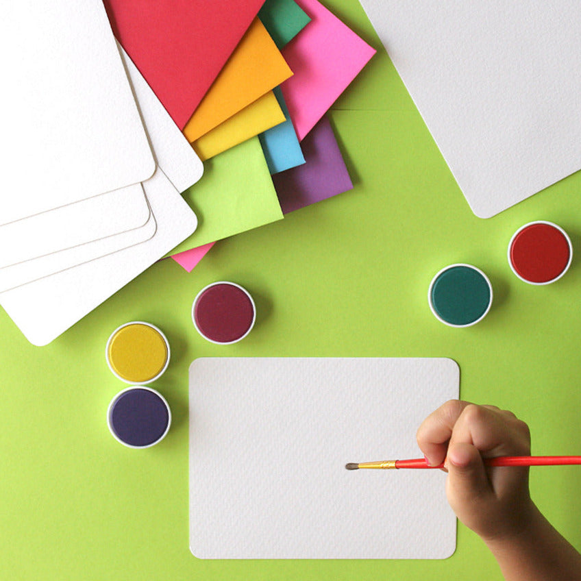 A person's hand is depicted at the bottom of the image, painting on a white watercolor paper flat card using a paintbrush. Surrounding the card are various colorful paints and envelopes, suggesting artistic activity.