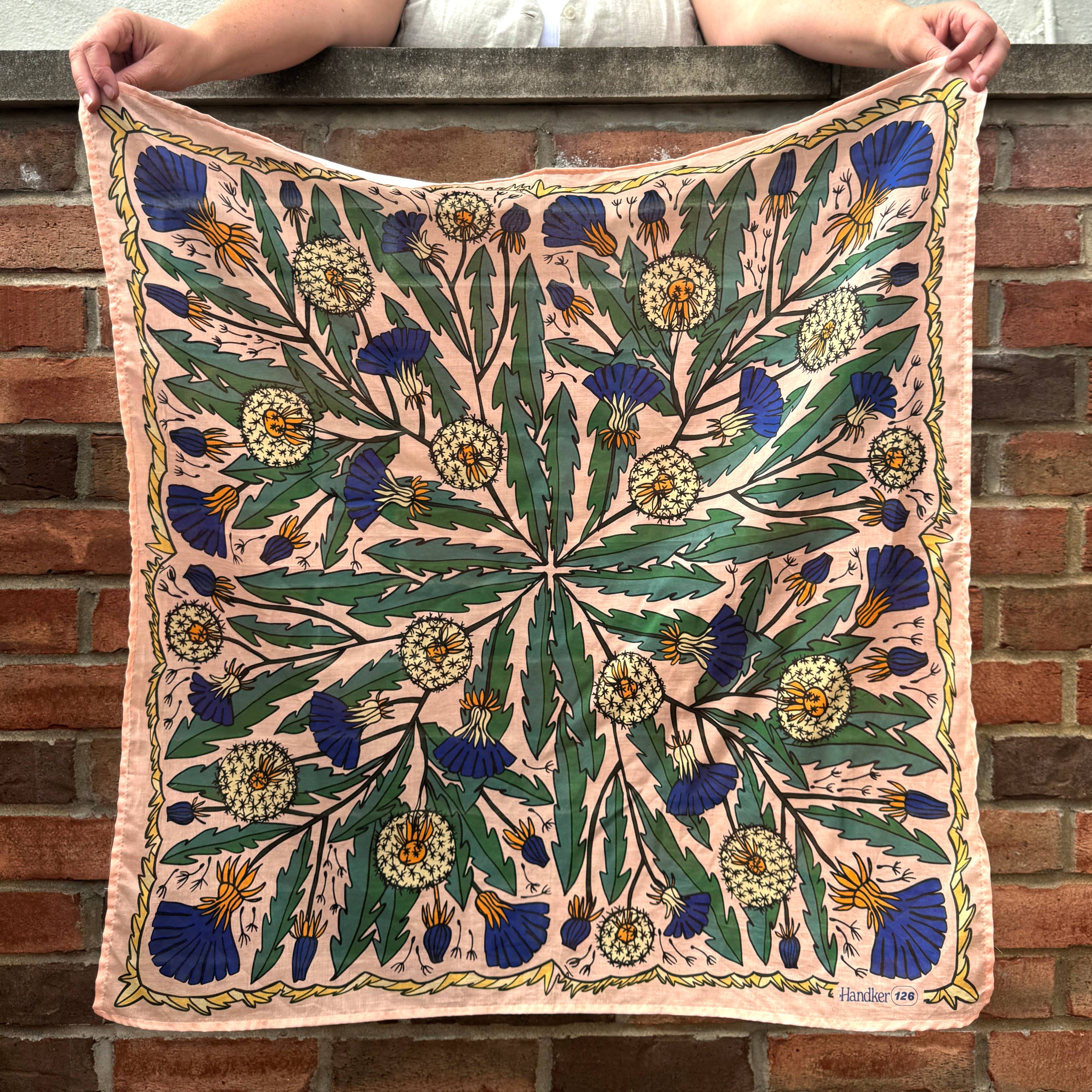 Person holding a square dandelion patterned wild rag scarf against a brick wall