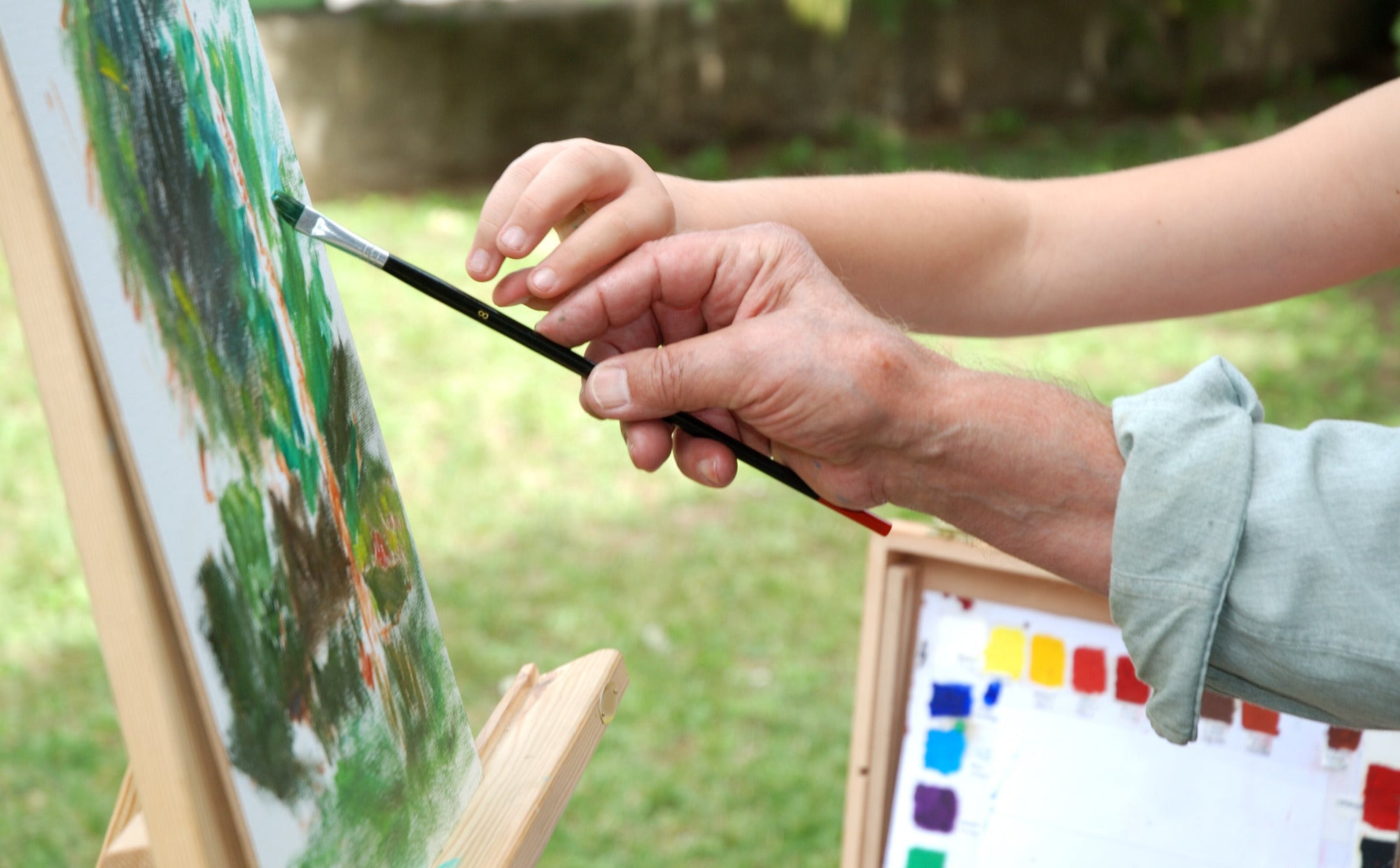Adult and child hands holding paintbrush and painting together outside with easel