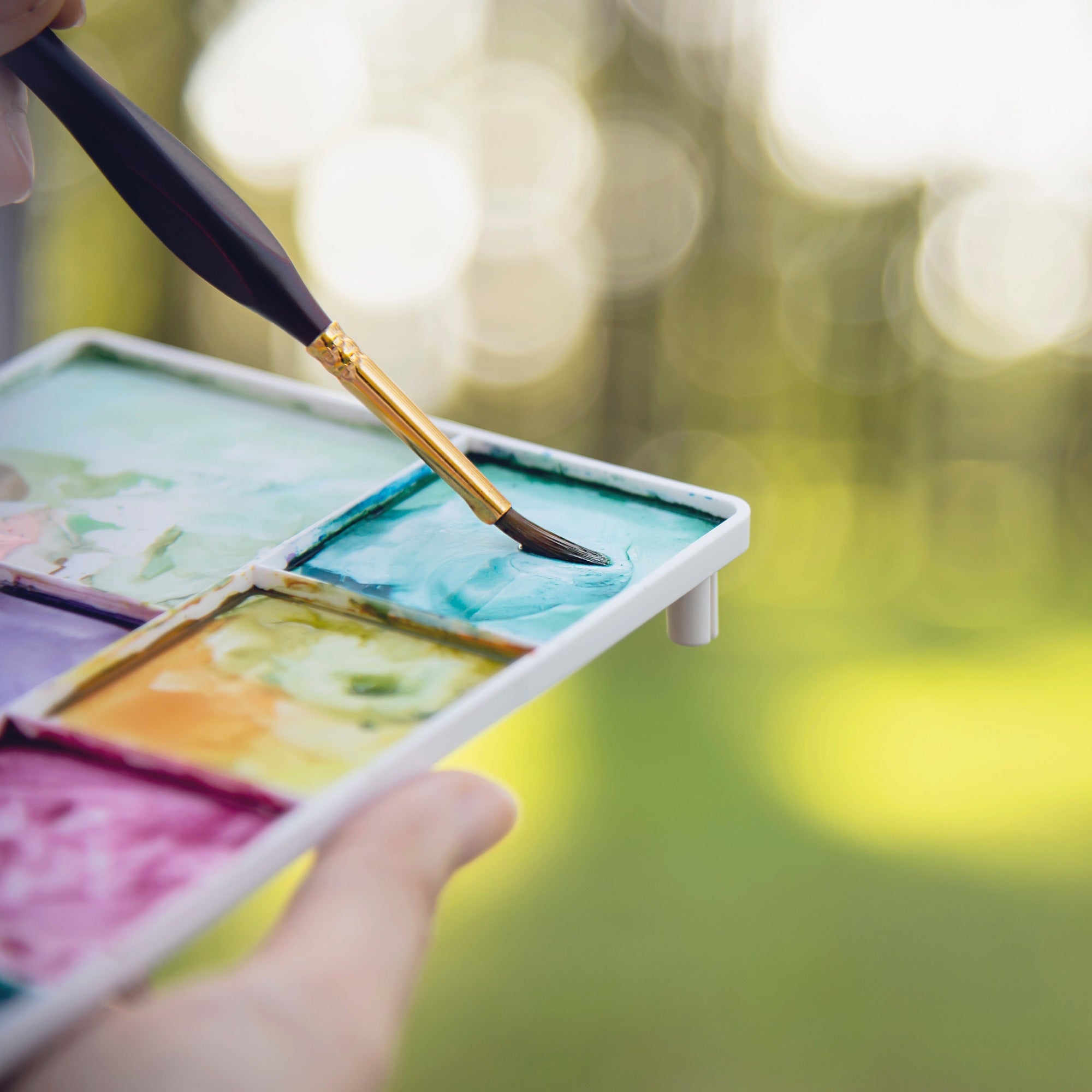 Close up of human hand holding a paint pallet and a paintbrush blurry outside background