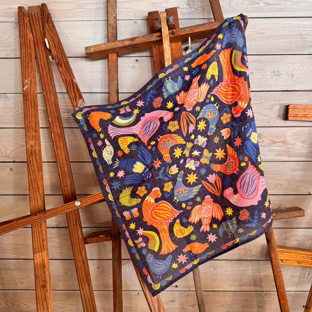 Colorful patterned scarf with chickens and birds draped over a wooden easel on a wooden floor.