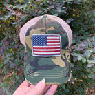 Camouflage cap with American flag patch held in front of a leafy background