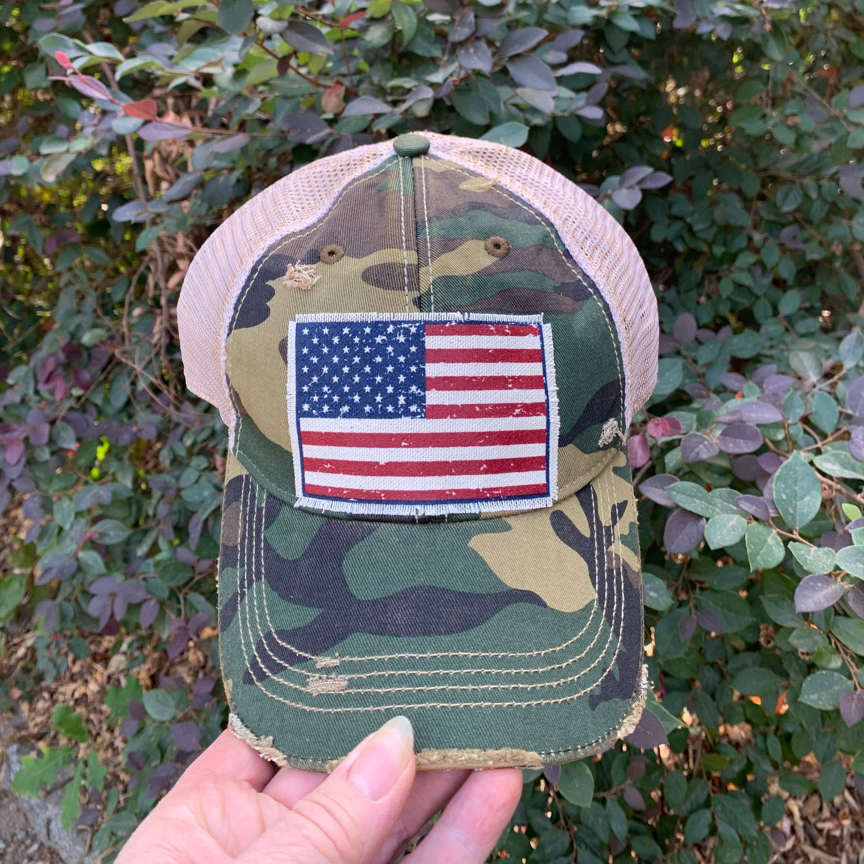 Camouflage cap with American flag patch held in front of a leafy background