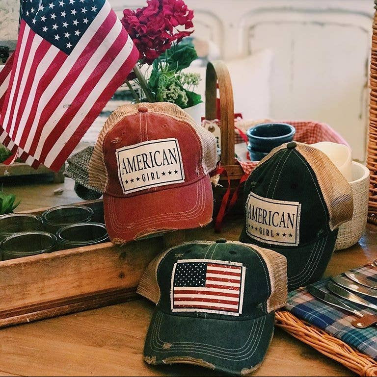Three hats with 'American Girl' and American flag designs on a table with a flag and flower plant in the background.