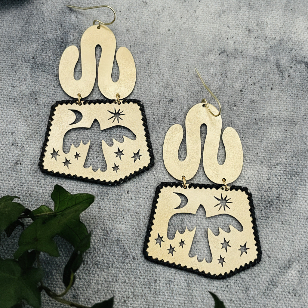 Pair of handmade brass earrings with cut-out designs of birds and celestial elements on a textured surface