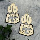 Pair of handmade brass earrings with cut-out designs of birds and celestial elements on a textured surface