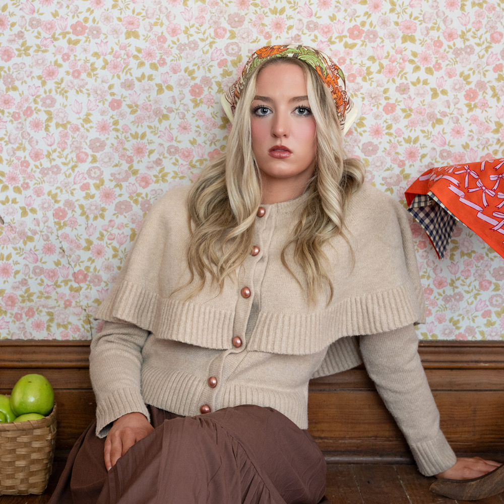 Woman sitting on a the floor with a floral patterned wall behind her, next to a basket of apples and wearing a floral cotton bandana as a head scarf