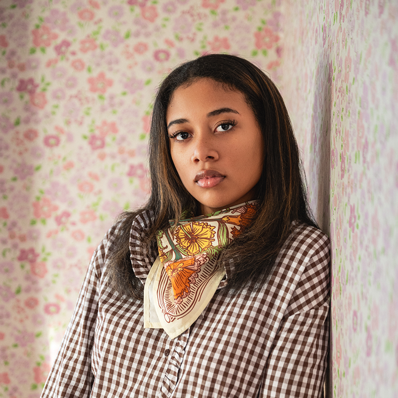 Woman wearing a checkered shirt with a floral patterned cotton bandana scarf against a floral wallpaper background