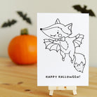 Halloween-themed card with a fox dressed as a vampire on a wooden surface with a pumpkin and bats in the background.
