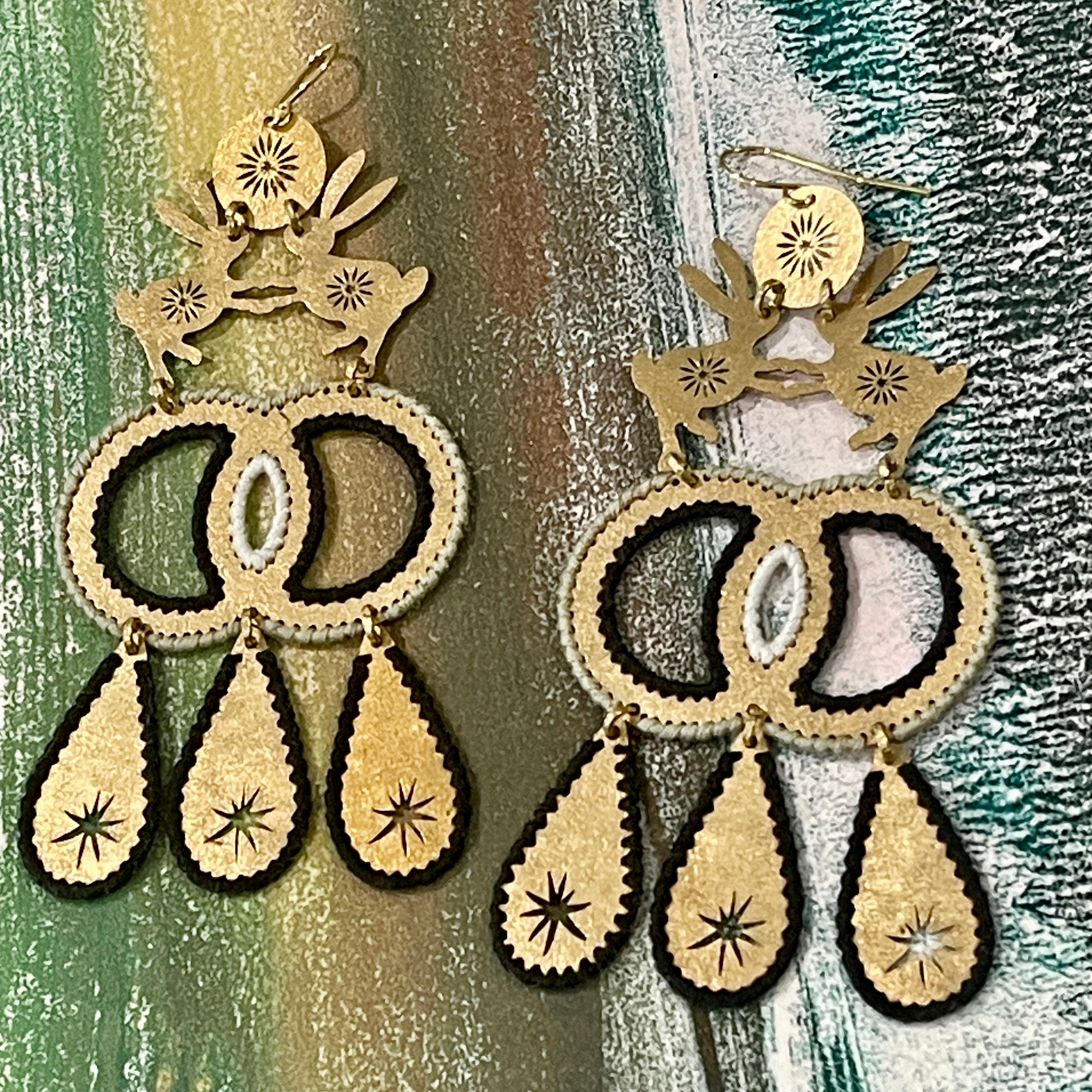 Pair of dangle earrings made from textured brass with two hares dancing and color thread edges on a muted color background