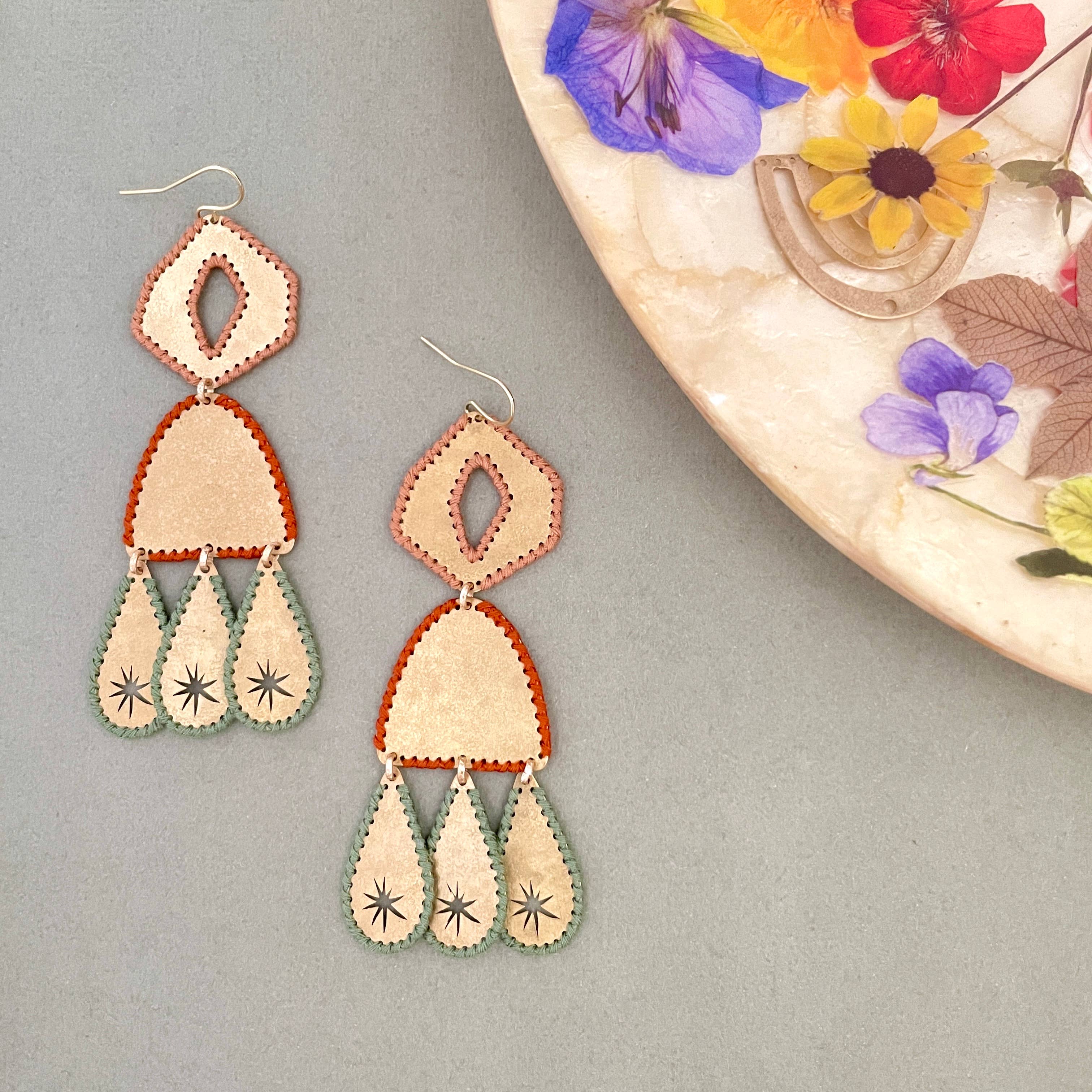 Pair of teardrop earrings made from brass with hand stitched thread edges next to a decorative plate on a gray background