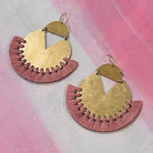 Pair of handmade brass earrings with coral color thread fringe detail on a pink painting background