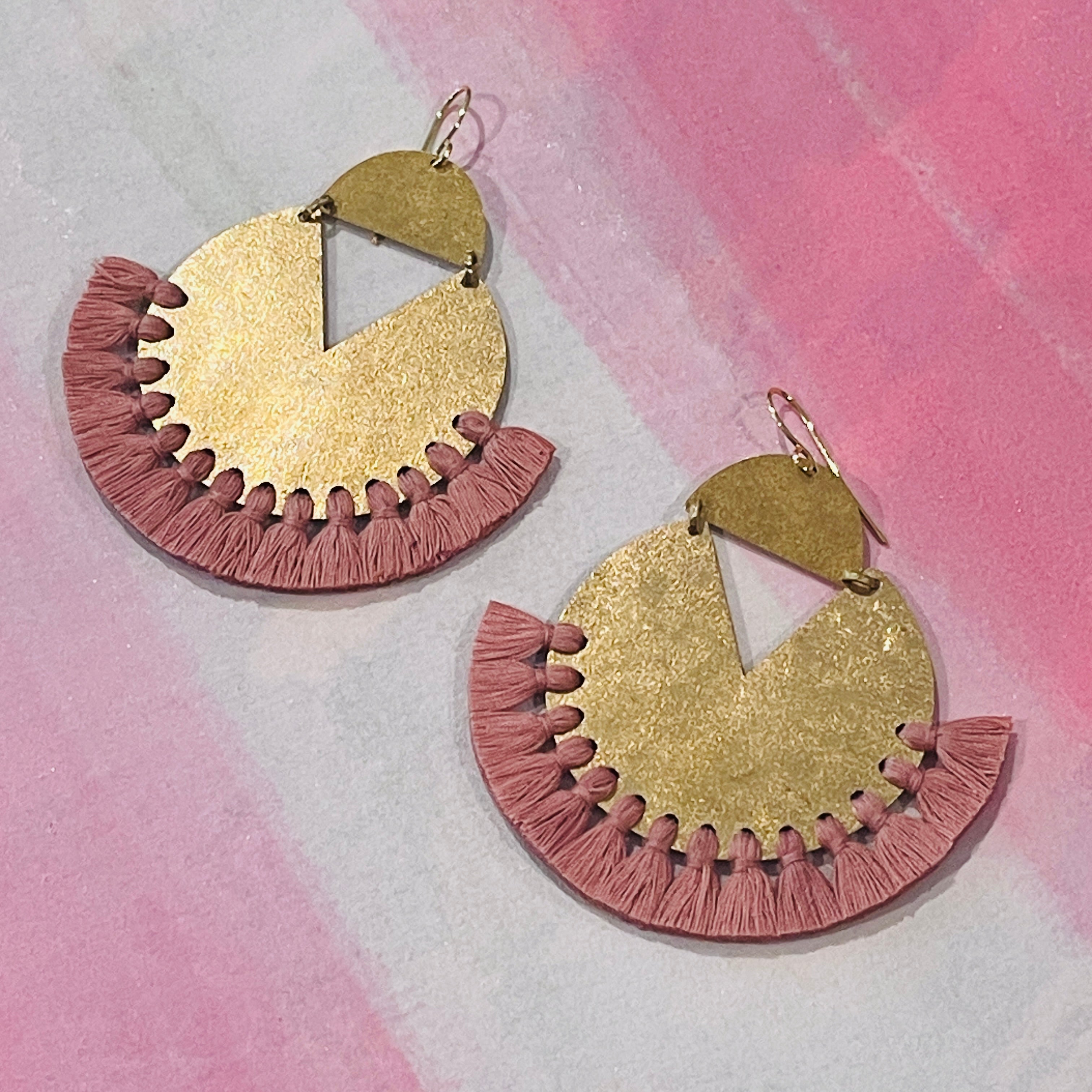 Pair of handmade brass earrings with coral color thread fringe detail on a pink painting background