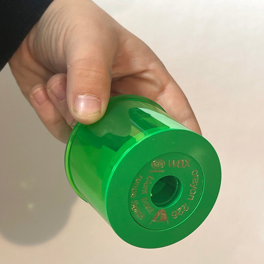 A child's hand holding a green jumbo crayon sharpener against a cream background