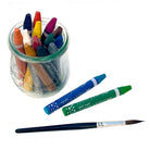 Set of 12 jumbo watercolor crayons in a glass storage jar with a green and a blue watercolor crayon and a paint brush laying next to the jar on white background