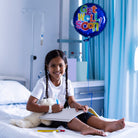 Child in a hospital room with a 'Get Well Soon' balloon, holding a teddy bear and drawing in a sketchbook.