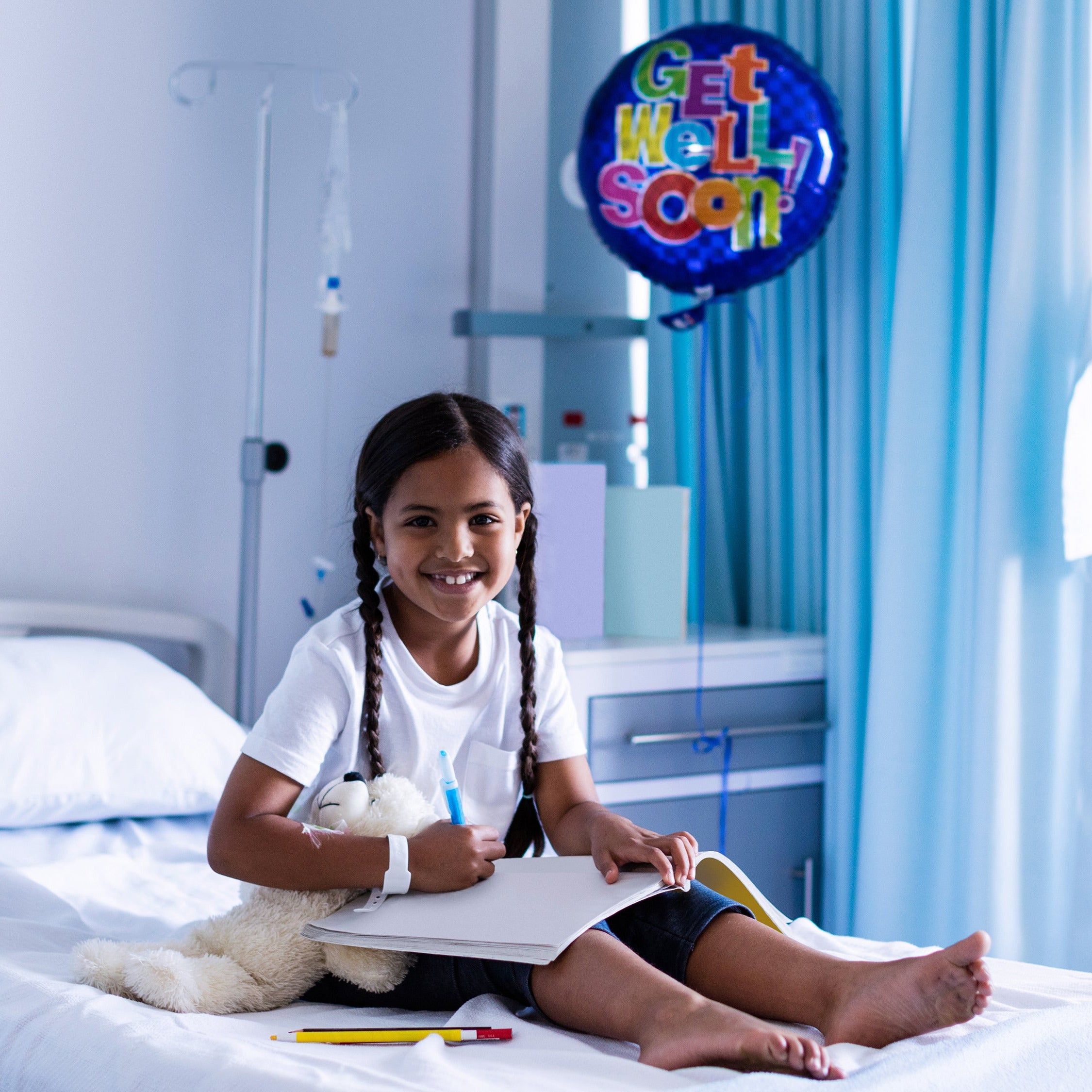 Child in a hospital room with a 'Get Well Soon' balloon, holding a teddy bear and drawing in a sketchbook.
