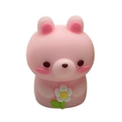 A small pink bunny shaped pencil sharpener with pink inner ears, dark brown eyes and nose, pink cheeks, and a white daisy flower with a green stem held in front of its chest, shown on a plain white background.