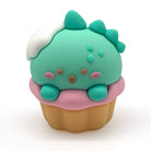 A small cupcake-shaped pencil sharpener with a light green dinosaur face and body, white frosting, and a pink frosting cupcake base, centered on a plain white background.