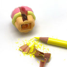 A close-up of a yellow pencil lying beside broken brown pencil shavings and a partially sharpened pencil tip, with a pink and yellow pencil sharpener visible in the background on a white surface.