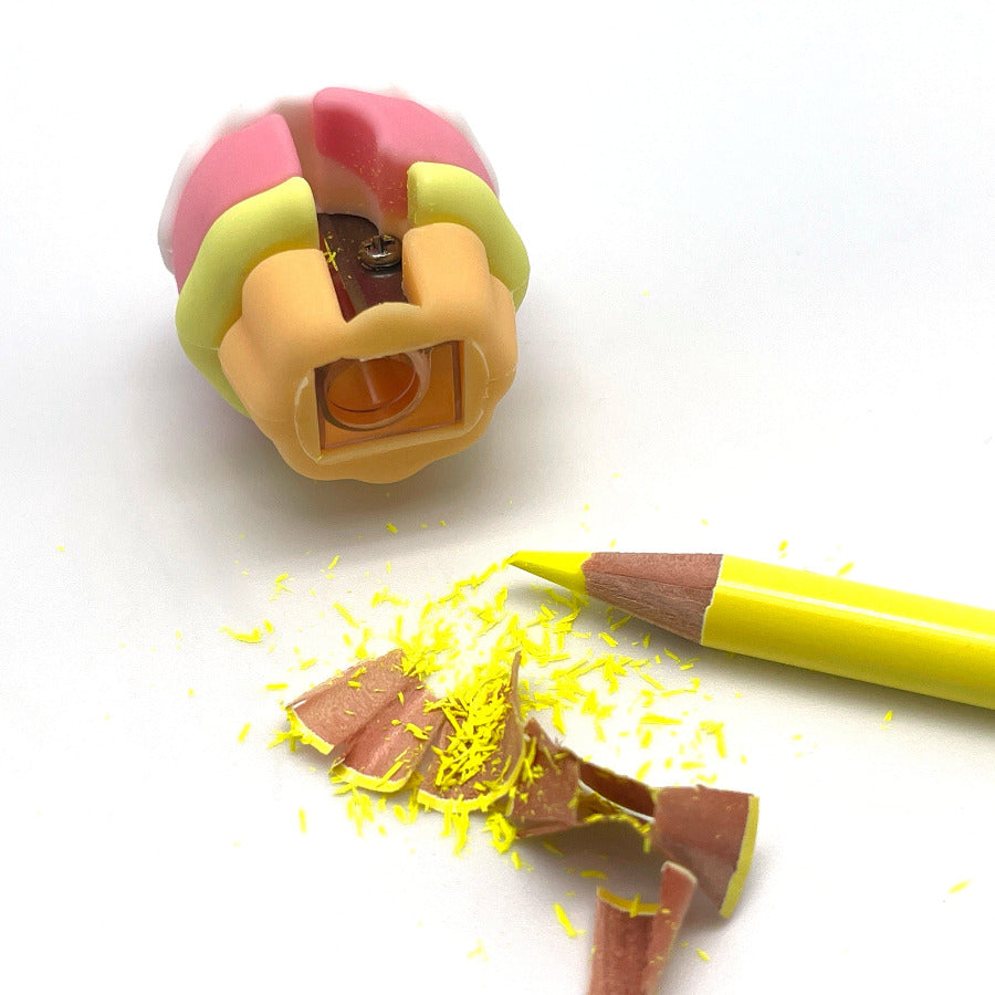 A close-up of a yellow pencil lying beside broken brown pencil shavings and a partially sharpened pencil tip, with a pink and yellow pencil sharpener visible in the background on a white surface.