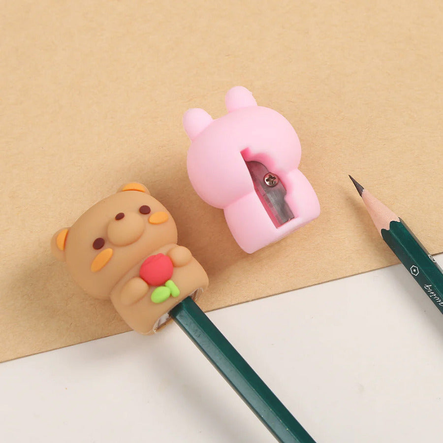 Two cute pencil sharpeners, one tan bear holding a red flower and one pink bunny, shown from behind with a green pencil on a beige and white background.