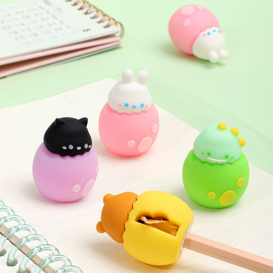 Cute colorful pencil sharpeners designed like baby animals hatching out of eggs on a mint green background with notebooks and a pencil being sharpened