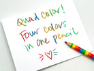 A white note card with colorful handwritten text reading 'Quad color! Four colors in one pencil' and a rainbow-striped colored pencil placed on the lower right, photographed on a light gray background.