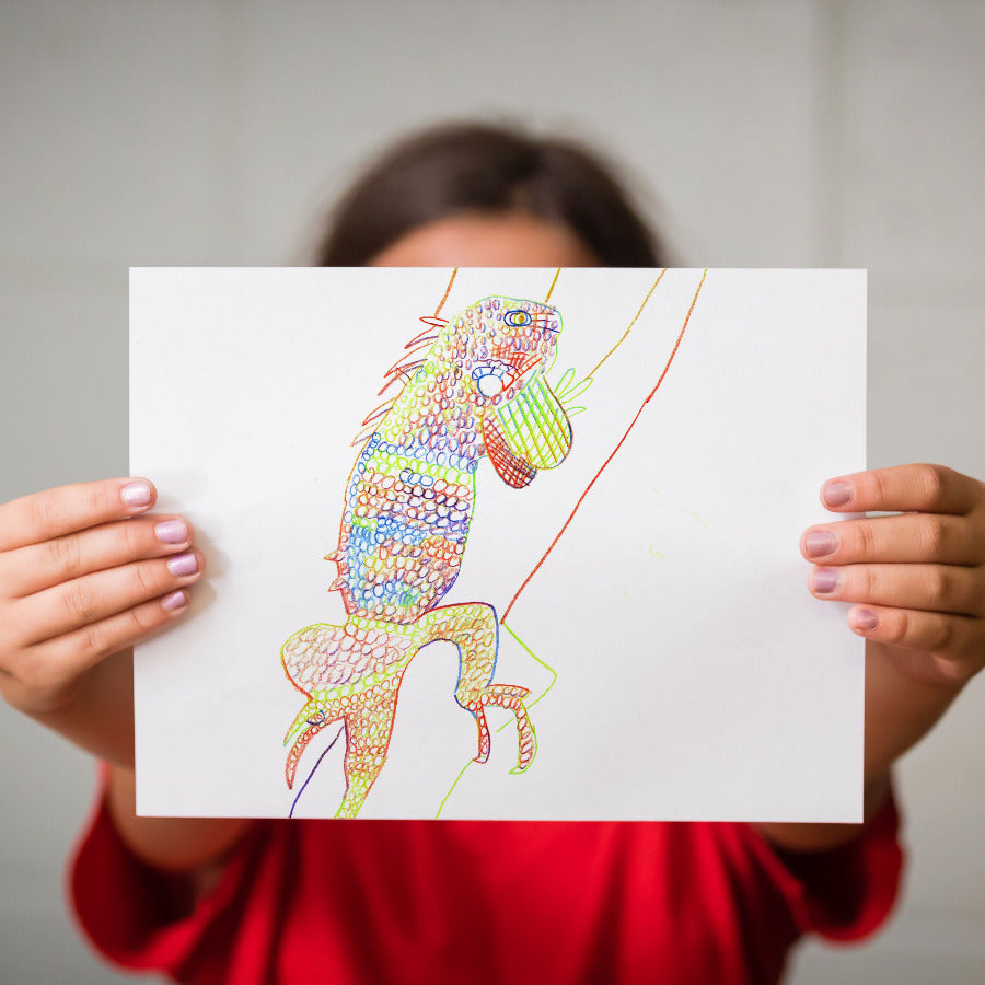 A child holds up a vivid, multi-color swirl drawing of a lizard drawn with a rainbow pencil. The child's hands, hair, and red shirt are visible against a neutral gray wall background.