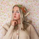 Woman wearing a beige cardigan and colorful floral cotton bandana as a headscarf against a floral wallpaper background