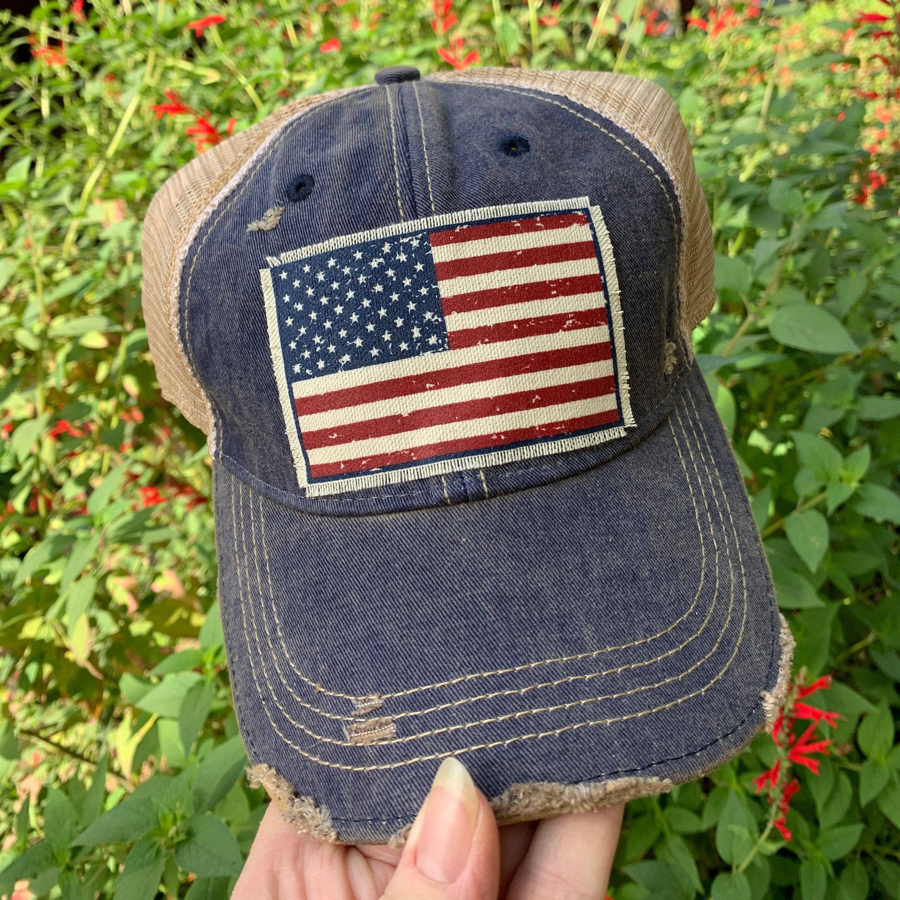 Blue baseball cap with American flag patch held in front of green foliage and red flowers