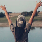 Person wearing a 'Country Girl' cap with arms raised by a body of water