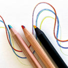 Three colored rainbow pencils on a white surface with colorful drawn lines behind them. The pencils include a black pencil, a natural wood pencil, and a triangle shape wood pencil with rainbow tips.