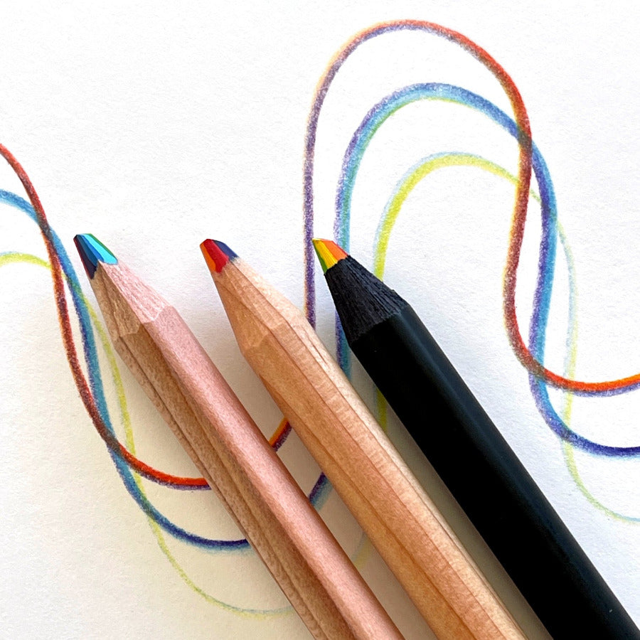 Three colored rainbow pencils on a white surface with colorful drawn lines behind them. The pencils include a black pencil, a natural wood pencil, and a triangle shape wood pencil with rainbow tips.